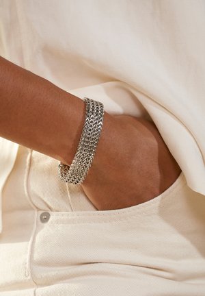 Silberne Kettenarmband mit verflochtenen Gliedern, das ein flaches, strukturiertes Design aufweist. Am Handgelenk getragen, kombiniert mit einem hellen Stoff.