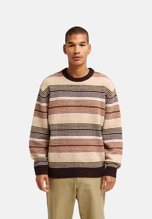Man draagt een beige, bruine en zwarte horizontaal gestreepte trui en lichtbruine broek, staand tegen een effen lichte achtergrond.