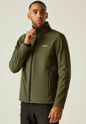 Mężczyzna w oliwkowo-zielonej kurtce softshell zapinanej na zamek z wysokim kołnierzem i czarnych spodniach, patrzący w lewo na jednolitym tle.