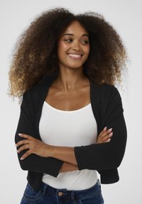 Eine Frau mit voluminösen, lockigen Haaren lächelt, trägt ein weißes Oberteil und eine schwarze Strickjacke, ihre Arme vor der Brust verschränkt, vor einem einfarbigen, hellen Hintergrund.
