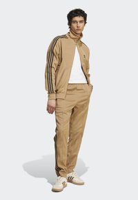 Tan tracksuit with black side stripes, zip-up jacket, and elastic waistband pants. Paired with white shoes featuring brown stripes.