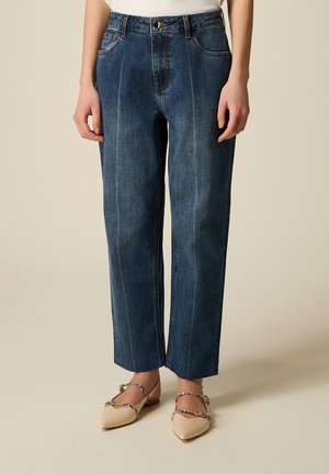 Jeans blu a gamba dritta con dettagli di cuciture frontali, indossati con una maglietta bianca e ballerine beige con punta a punta e doppi cinturini alla caviglia.