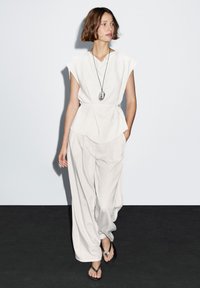 Lichtbeige mouwloze top met een uitlopende zoom, gecombineerd met loszittende lichtgrijze broek en zwarte slippers, tegen een neutrale achtergrond.