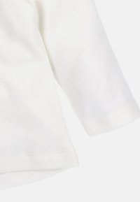 Biała koszula z długim rękawem wykonana z miękkiej bawełnianej tkaniny, z widocznym przeszyciem na mankiecie i dole, na jasnym tle.