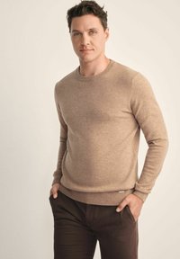 Be�żowy sweter dzianinowy z długimi rękawami, okrągłym dekoltem i subtelną strukturą, w połączeniu z ciemnobrązowymi spodniami i markowym akcentem na dolnym brzegu.