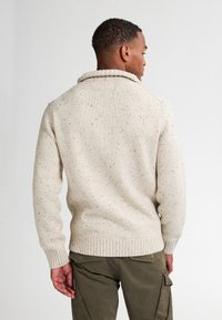 Maglione lavorato a maglia color crema con macchie testurizzate, colletto alto e orlo a coste. Indossato con pantaloni cargo verde oliva, visto da dietro.