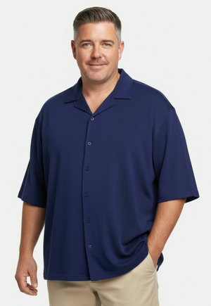 Mann mit kurzen Haaren, der ein marineblaues kurzärmeliges Hemd mit Knöpfen und beige Hosen trägt, steht mit einer Hand in der Tasche.
