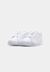 Białe sneakersy wykonane z gładkiego materiału, z niskim krojem, sznurowane, z płaską gumową podeszwą i subtelnym logo po boku.