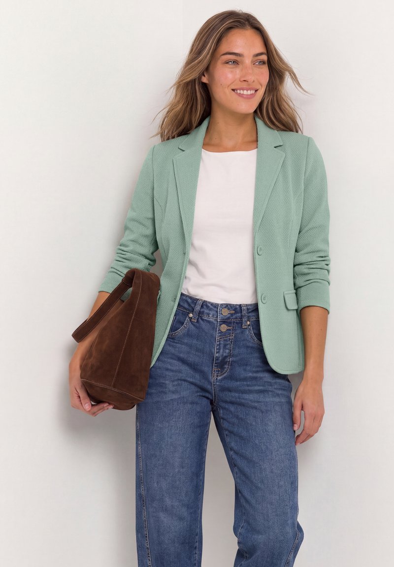 Femme portant un blazer vert clair, une chemise blanche et un jean bleu, tenant un sac à main en daim marron, souriante et regardant sur le côté.