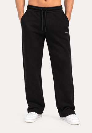 Hombre con pantalones deportivos negros sueltos con cordón y zapatillas blancas, con las manos en los bolsillos, de pie contra un fondo liso.