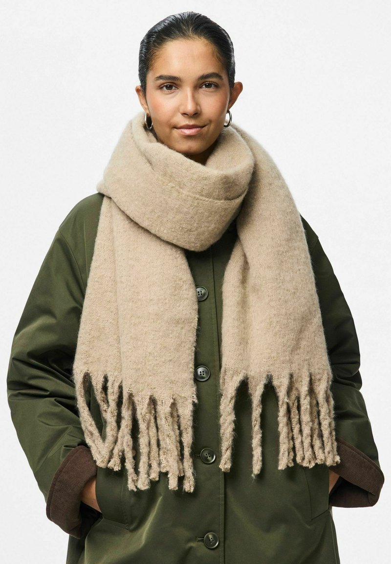 Sciarpa di lana beige con estremità frange, indossata sopra un cappotto verde oliva. La sciarpa è spessa, testurizzata e si avvolge liberamente attorno al collo.
