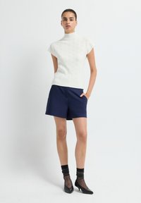 White, short-sleeve knitted top with a high collar, paired with navy shorts. The model wears black heels and textured black socks.