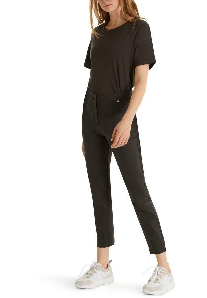 Femme portant un haut noir à manches courtes, un pantalon noir ajusté longueur cheville et des baskets blanches, debout avec une main détendue à ses côtés.