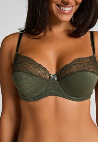 Olijfgroene bh met kantdetails aan de bovenkant, een gepunte textuur op de cups en een klein grijs strikje in het midden van het gore. Verstelbare bandjes.