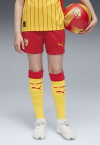 Conjunto de fútbol de Puma que incluye una camiseta de rayas amarillas, pantalones cortos rojos, calcetas amarillas con acentos rojos y zapatillas blancas, sosteniendo un balón de fútbol rojo.