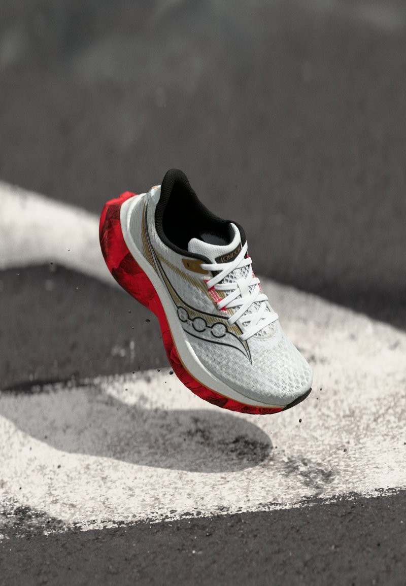 Λευκό και κόκκινο αθλητικό παπούτσι με μαύρες και χρυσές λεπτομέρειες, στον αέρα πάνω από μια υφή γκρι επιφάνεια με λευκές γραμμές.