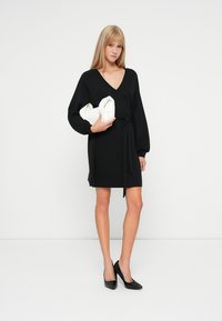 Vestido negro de manga larga con escote en V y detalle de cinturón, combinado con un bolso blanco de mano y tacones negros de punta. Tela suave y diseño ajustado.