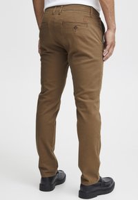Pantalones de algodón marrones con corte ajustado, que cuentan con dos bolsillos traseros con botones y un parche con el logotipo, combinados con zapatos negros.