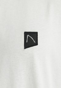 La maglietta in tessuto grigio chiaro presenta una toppa quadrata nera con un design geometrico bianco. Il materiale appare morbido e con una texture liscia.