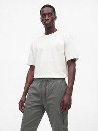Hombre con camiseta blanca lisa de manga corta y pantalones cargo grises de pie frente a un fondo blanco.