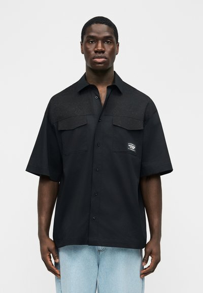 Mladý muž v krátkém černém košili s kapsami na hrudi a světlých modrých džínách stojí před jednoduchým pozadím.