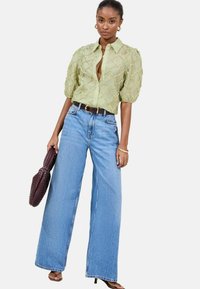 Hellgrüne, strukturierte Bluse mit Puffärmel und Knopfverschluss, kombiniert mit hochgeschnittenen, weit geschnittenen blauen Jeans. Accessoires: Eine dunkle Handtasche.