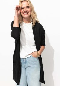 Cardigan noir à ouverture frontale, fabriqué en tissu doux. Porté sur un t-shirt blanc et associé à un jean bleu clair. Dispose d'une coupe décontractée et de poches.