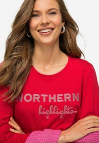 Červená košile s dlouhým rukávem, na které je šedý text "NORTHERN highlights", kulatý výstřih, měkká tkanina, nošena s růžovým pleteným doplňkem.