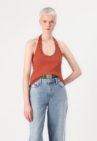 Haut halter de couleur rouille avec un col rond et des œillets en métal argenté. Associé à un jean taille haute bleu clair. Fond uni.