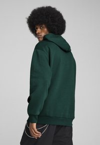 Tamsiai žaliai džemperis su reguliuojama kapuce, raugintais rankogaliais ir apatine dalimi, bei laisvu pjaustymu. Galinėje kišenėje yra sidabrinės grandinės akcentas.