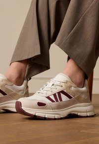 Sneakers bianchi e beige con accenti color bordeaux, sovrapposizioni testurizzate, design da scarpa da corsa e suola ammortizzata, abbinate a pantaloni marroni.