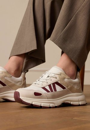 Witte en beige sneakers met bordeaux accenten, gestructureerde overlays, ontwerp van hardloopschoenen en een gevoerde zool, gecombineerd met bruine broek.