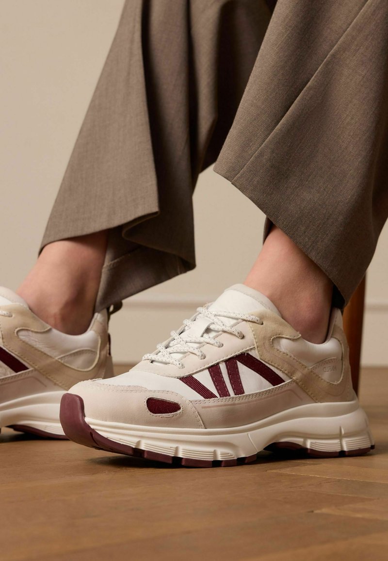 Sneakers bianchi e beige con accenti color bordeaux, sovrapposizioni testurizzate, design da scarpa da corsa e suola ammortizzata, abbinate a pantaloni marroni.