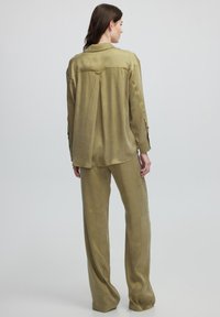 Camisa de lino verde oliva con un ajuste relajado, detalle de pliegue en la parte posterior, mangas largas y puños abotonados, combinada con pantalones anchos a juego.