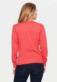 Pulover coral cu mâneci lungi, guler și tiv cu nervuri; textură netedă; design ajustat; purtat cu blugi negri.