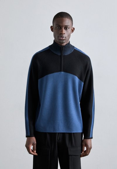 Pull-over zippé bleu et noir avec col montant, blocs de couleurs contrastants, texture lisse et poignets côtelés.