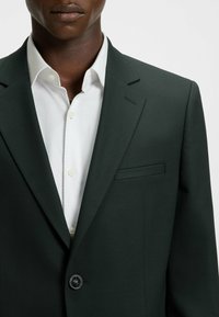 Dunkelgrüner Blazer mit strukturiertem Finish, einreihiger Knopfverschluss und Brusttasche, kombiniert mit einem weißen Hemd im Fischgrätenmuster.