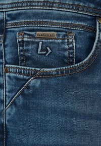 Granatowe dżinsy z tylną kieszenią z wyszytym logo 'L' oraz mosiężnymi akcentami. Materiał ma klasyczną fakturę i wykończenie.