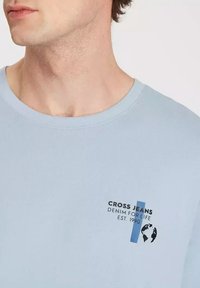 Jasnoniebieska bawełniana koszulka z krótkim rękawem, z graficznym nadrukiem zawierającym napis "CROSS JEANS" oraz emblematem globu na przodzie.