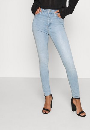 Femme portant un jean skinny bleu clair et des sandales à talons hauts noires ouvertes à l’avant, mains dans les poches, debout contre un fond blanc uni.