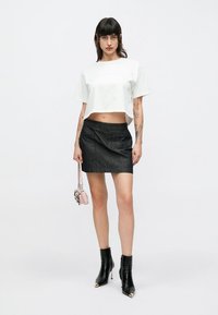 T-shirt bianca corta, minigonna in denim scuro, stivaletti neri con dettagli dorati, che tiene una piccola borsa rosa. Tessuto liscio, design aderente.