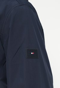 Tengerészkék dzseki ujja fekete, téglalap alakú Tommy Hilfiger logóval, amely piros, fehér és kék zászló mintázatot tartalmaz.