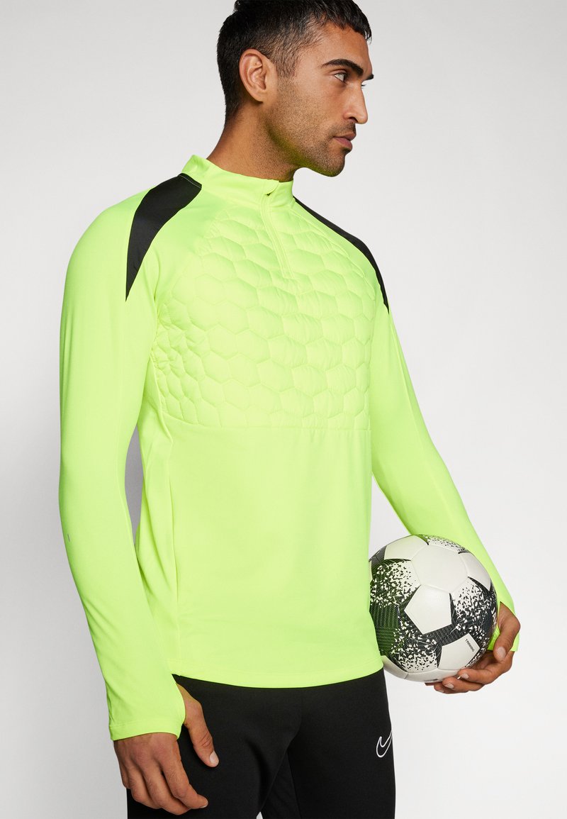 Maglietta atletica a maniche lunghe verde neon con motivo a rete esagonale sul petto e dettagli neri sulle spalle, abbinata a un pallone nero.
