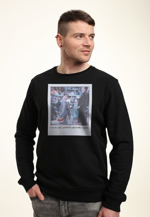 Homme aux cheveux courts portant un sweat-shirt noir avec un graphisme coloré de deux hommes dans une cuisine et le texte « Could I be wearing anymore clothes ».