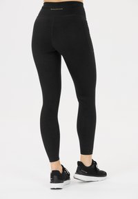 Persoon in zwarte high-waist legging met subtiel patroon en "Endurance" label, gecombineerd met zwart-witte sport schoenen op een witte achtergrond.