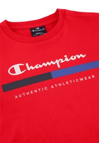 Camiseta de algodón roja con cuello redondo, que presenta el logo de "Champion" en blanco y una franja horizontal azul y negra con el texto "AUTHENTIC ATHLETICWEAR".