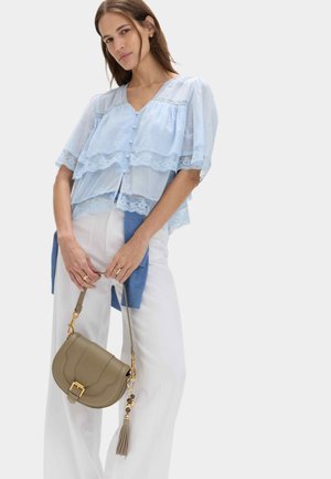 Kvinde iført lagdelt lyseblå bluse med blonder, hvide bukser, der holder en beige håndtaske med frynser og gulddetaljer mod en ensfarvet baggrund.