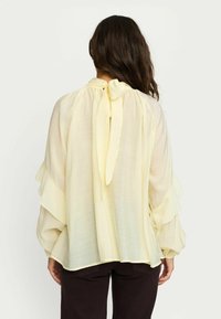 Blouse jaune légère avec des manches à volants et un lien dans le dos. Texture transparente et coupe décontractée. Associée à un pantalon marron foncé.