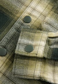 Gros plan sur un tissu à carreaux vert et beige avec des boutons-pression vert foncé sur le poignet et la patte de boutonnage avant de la chemise.