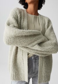 Lichtgroene, getextureerde cardigan met een relaxte pasvorm en open voorkant, gelaagd over een geribbeld, lichtbeige top. Zacht, pluizig materiaal met brede mouwen.